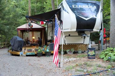 Fifth wheel RV with covered patio and seating area at a wooded campground