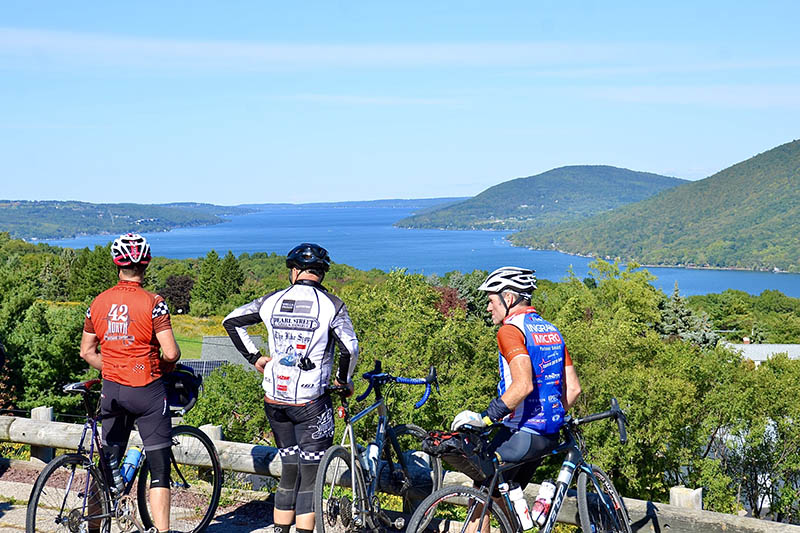 Three cyclists enjoying a scenic overlook with a Finger Lake and forested hills in the background.