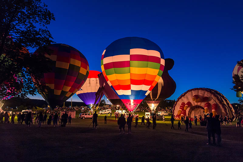 Colorful hot air balloons illuminated at dusk during an outdoor festival with visitors gathered nearby.